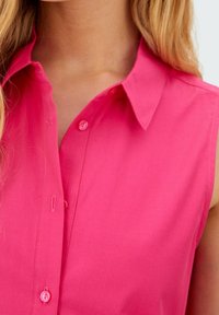 Ilman hihoja oleva napitettava paita kirkkaan vaaleanpunaisesta kankaasta, jossa on kauluskansio ja yksittäinen nappirivi edessä. Sileä rakenne.