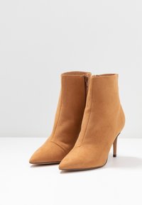 Paire de bottines en daim beige avec bouts pointus, fermetures éclair latérales et talons hauts fins sur une surface blanche avec un fond uni.