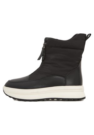 Bottes d'hiver montantes noires avec fermetures éclair à l'avant et semelles blanches épaisses, portées avec des leggings noirs et des guêtres blanches en tricot amples.