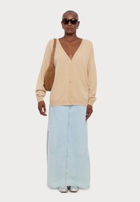 Beżowy sweter z dzianiny z dekoltem w serek, w połączeniu z jasnoniebieską jeansową spódnicą maxi z bocznymi rozcięciami. Srebrne buty i brązowa torba na ramię.
