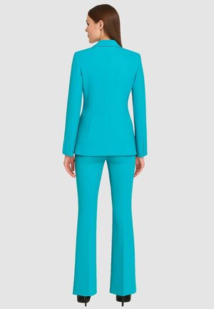 Donna con lunghi capelli castani in piedi di spalle, indossa un completo pantalone aderente di un brillante turchese con tacchi alti neri.