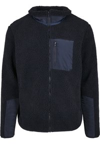 Veste en polaire bleu marine foncé avec fermeture éclair intégrale à l'avant, poche poitrine zippée et renforts en tissu sur les coudes et la poche.