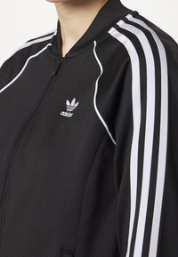 Svart zip-up jacka av slät tyg, med vita tre-randiga detaljer på axlarna och en liten vit logotyp på bröstet.