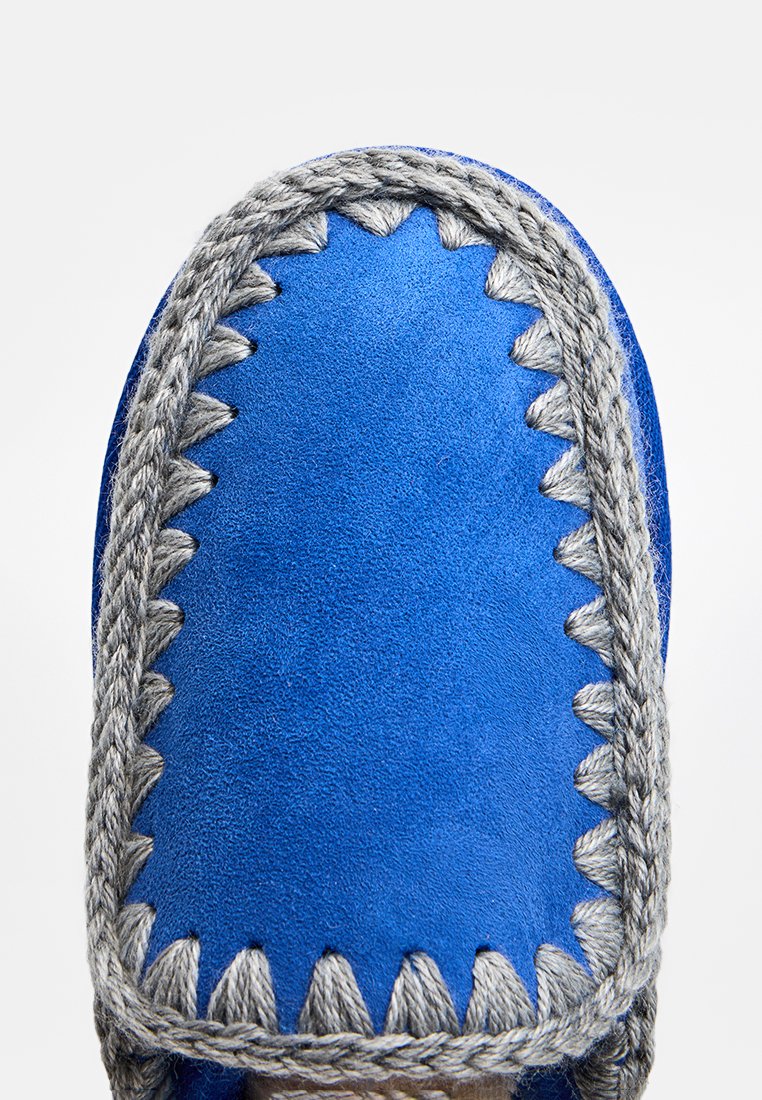 Mocassini di pelle scamosciata blu con spessa cucitura grigia intrecciata lungo la punta arrotondata e i bordi, su uno sfondo bianco.
