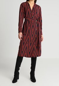 Abito avvolgente a zigzag rosso e nero con scollo a V, maniche lunghe e dettagli con bottoni, abbinato a stivali neri al ginocchio.