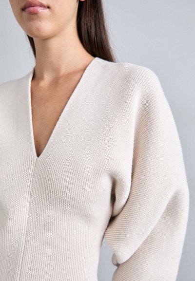 Gaiši bežs adīts džemperis ar dziļu V veida kaklu, ribota tekstūra un pūkaini piedurknes. Mīksta, elastīga auduma struktūra izceļ ķermeņa formu.