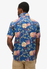 Korte mouw shirt van blauwe stof met een gedetailleerd patroon van oranje en witte bloemen, cirkelvormige motieven en gestileerde bogen.