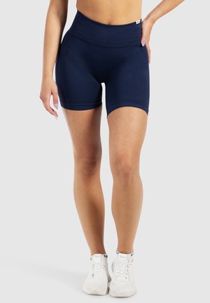 Kvinde iført marineblå atletikshorts med høj talje og hvide sneakers, stående med den ene hånd på hoften mod en ensfarvet baggrund.