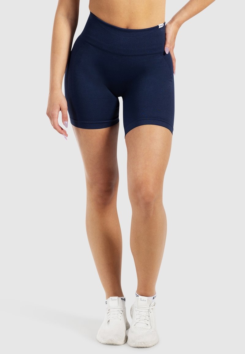 Femme portant un short de sport taille haute bleu marine et des baskets blanches, debout avec une main sur la hanche devant un fond uni.