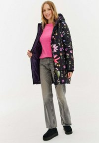 Černá prošívaná bunda s květinovým vzorem, lehká, s kapucí, v kombinaci s růžovým svetrem a šedými džínami, nošená s černými slip-on botami.