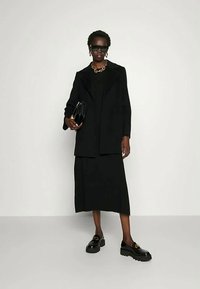 Schwarze taillierte Blazer über einem schwarzen Oberteil und einem Midikleid. Voluminöse schwarze Schuhe, große Sonnenbrille und eine goldene Kettenhalskette vervollständigen das Outfit.