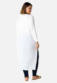 Lång vit cardigan med en texturerad tyg, med långa ärmar och sidoklyftor, i kombination med mörka byxor och platta sandaler.