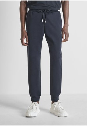 Uomo che indossa pantaloni sportivi blu navy con vita a coulisse e sneakers bianche, in piedi contro uno sfondo grigio uniforme.
