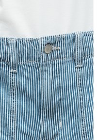 Gros plan sur un pantalon en denim à rayures verticales bleues et blanches avec un bouton en métal et des passants de ceinture sur un fond de tissu blanc.