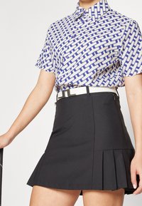 Košile s krátkým rukávem s modro-bílým geometrickým vzorem, spárovaná s černou skládací sukní a bílým pásem. Jednoduché pozadí.