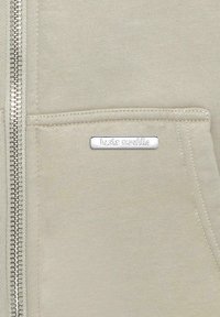 Lys beige zip-opp hettejakke laget av myk stoff, med en frontlomme og en sølvlogo som er sydd på lommen. Glidelås går nedover fronten.