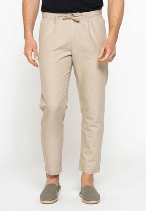 Uomo che indossa pantaloni beige con coulisse e scarpe verdi slip-on in piedi davanti a uno sfondo bianco.