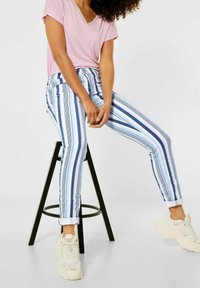 Randiga denimjeans med blå och vita vertikala linjer, kombinerade med en rosa V-ringad t-shirt och ljusa sneakers, sittande på en svart pall.