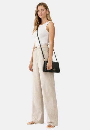 Donna con top bianco senza maniche, pantaloni beige ampi, sandali bianchi e una borsa a tracolla nera con un logo dorato.