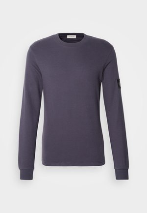 Chemise à manches longues violet foncé en tissu côtelé. Elle présente un col rond et un petit logo brodé sur la manche gauche.
