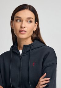 Hoodie nera con zip e coulisse, con un piccolo logo rosso sul lato sinistro del petto. Il materiale sembra morbido e testurizzato.