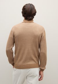 Bruine geribbelde trui met een hoge hals en een gestructureerde afwerking. De manchetten en zoom zijn ook geribd. Gecombineerd met lichtgekleurde broek.