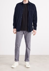 Tmavě modrá kordurová košile na knoflíky, nošená přes černé tričko, v kombinaci se světle šedými vyhrnutými kalhotami a bílými teniskami. Jednoduchý design a textura.