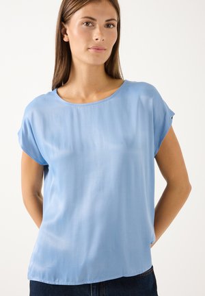 Frau mit glatten braunen Haaren, die eine hellblaue kurzärmelige Bluse und dunkle Hosen vor einem einfarbigen Hintergrund trägt.
