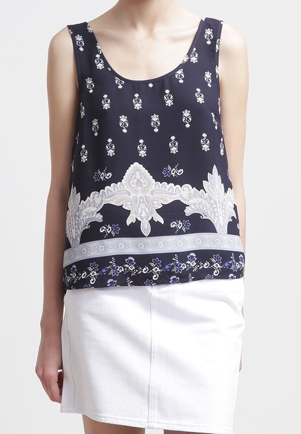 Femme portant un haut bleu marine sans manches avec des motifs floraux blancs et une jupe blanche, debout devant un fond uni.