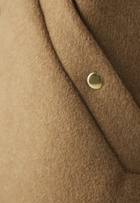 Brūns samts ar mīkstu tekstūru, kuram ir izteikta zelta pogu detaļa un diagonālu šuvju dizains.