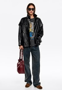 Čierna kožená nadrozmerná bunda, grafické tričko, široké denimmové rifle, hnedé slip-on topánky a bordová kožená dámska taška so zacvakávacími detailmi.