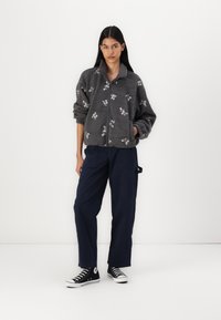 Casaco de fleece cinza com fecho zip, decorado com bordado floral, combinado com calças largas azuis marinhas e tênis de lona pretos com solas brancas.