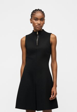 Ženska z pletenimi lasmi, oblečena v črno obleko brez rokavov z zadrgo spredaj in A-črtasto krilo, stoji pred enostavno svetlo ozadje.