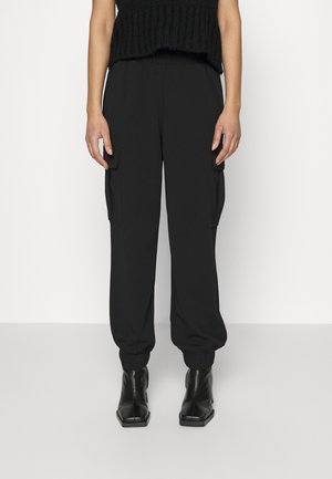 Person iført sorte cargo bukser med elastiske manchetter og sorte ankelstøvler, stående mod en ensartet lys baggrund.