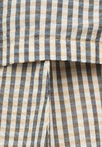 Ύφασμα σε μπεζ, μαύρο και λευκό με καρό σχέδιο, λεπτομερή ραφές και κεντρική πτυχή ή δίπλωμα.