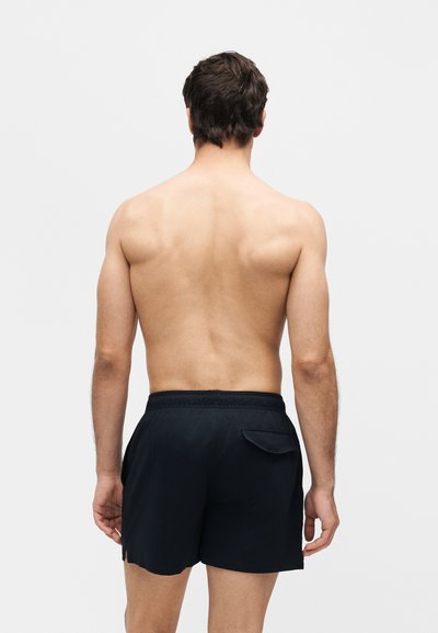 Άνδρας στέκεται με την πλάτη προς τα έξω, χωρίς πουκάμισο, φοράει μαύρο μαγιό με πίσω τσέπη μπροστά σε ένα απλό φωτεινό φόντο.