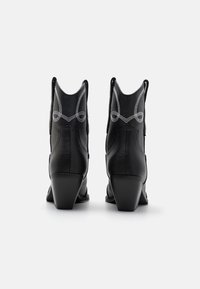Par črnih usnjenih kavbojkinih škornjev z belo dekorativno vezenino, gledano od zadaj na enobarvni beli podlagi.