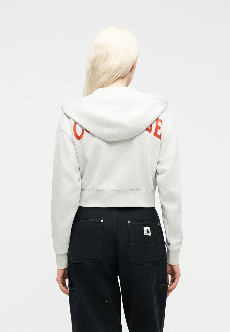 Vrouw met lang blond haar, gekleed in een lichtgrijze capuchontrui met rode tekst op de achterkant en zwarte cargobroek, tegen een effen achtergrond.