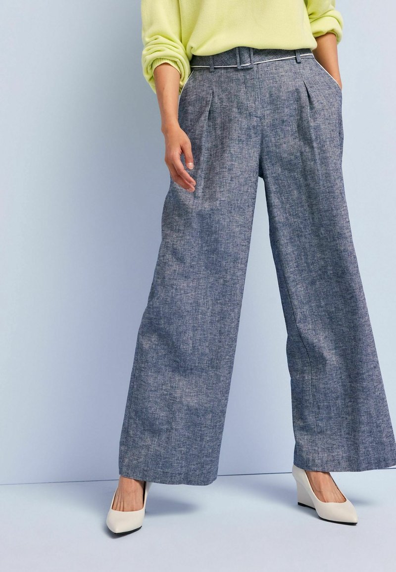 Personne portant un pull jaune clair, un pantalon gris à jambes larges avec des plis à l'avant et des chaussures compensées blanches à bout pointu, debout sur un sol clair.