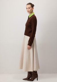 Brūns adīts kardigāns ar dimanta rakstiem, zaļa krekla apkakle, krēmkrāsas A-veida svārki, brūnas augstpapēžu zābaki. Gludas tekstūras, strukturēta silueta.