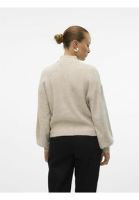 Un pull en maille côtelée beige clair avec un col montant et des manches ballon. Le bas est légèrement effiloché. Associé à un jean noir.
