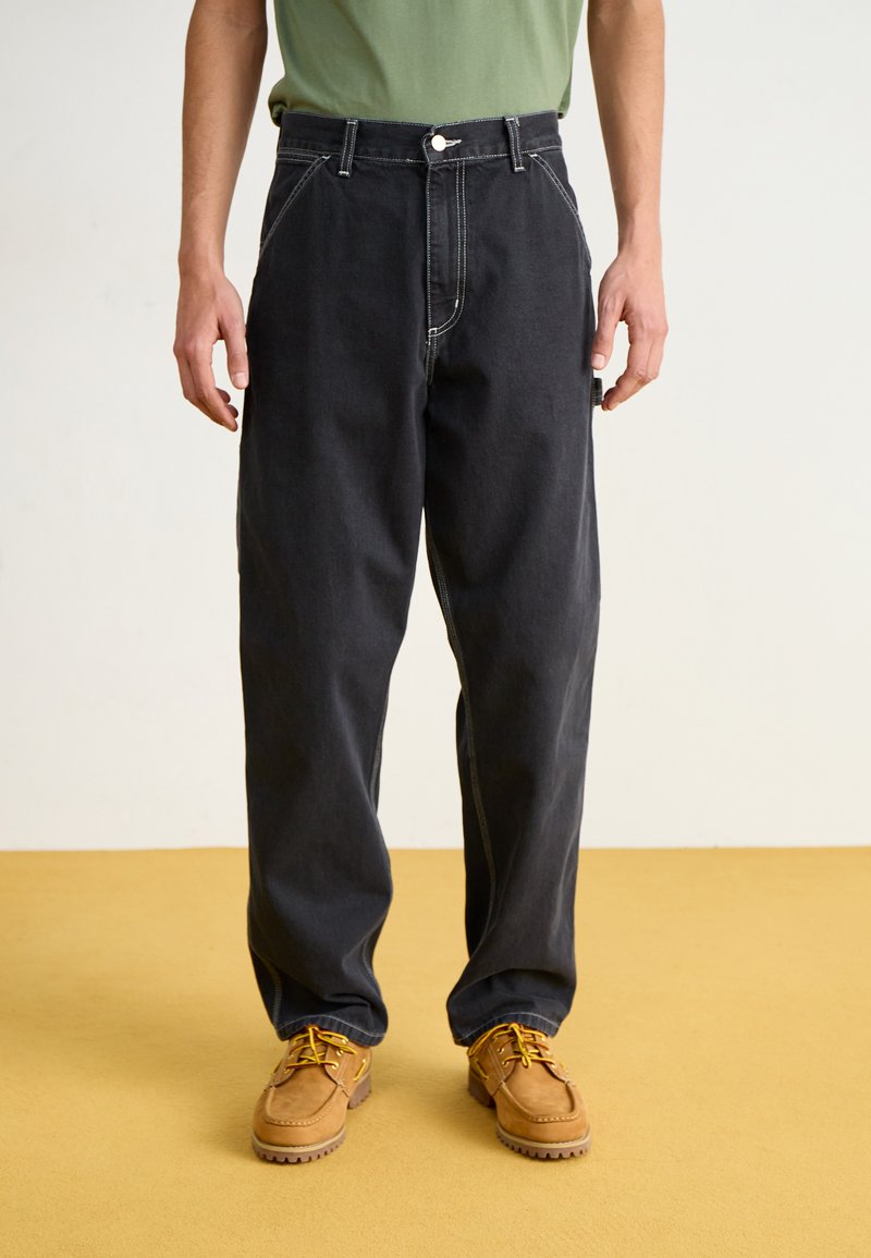 Dark denim trousers featuring a high waist, side pockets, contrasting stitching, and a relaxed fit, paired with tan lace-up shoes.