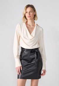 Kremfarget bluse med drapert design og lange ermer, sammen med et svart faux skinnskjørt med knytting i midjen.