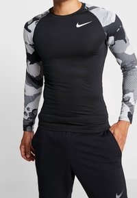 Długi rękaw koszulka sportowa, czarny korpus z rękawami w szarym wzorze moro, wykonana z elastycznego materiału, z logo Nike na piersi.