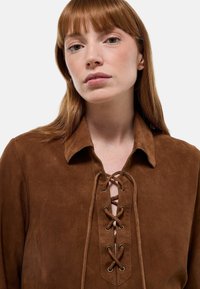 Blouse en daim marron avec col, devant lacé et texture lisse. Comprend des œillets en métal et une coupe décontractée.