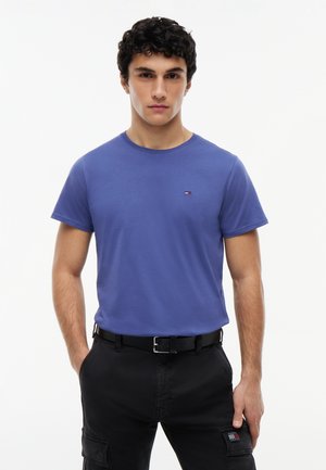 Mann mit blauem Rundhals-T-Shirt und schwarzen Cargohosen mit Händen in den Taschen, stehend vor einem schlichten weißen Hintergrund.