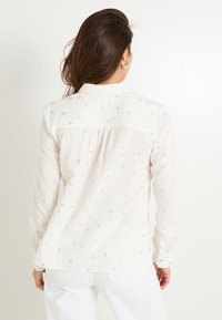 Mujer con cabello largo y castaño, vestida con una blusa blanca con pequeños patrones de lazo dorado y pantalones blancos, de espaldas contra un fondo liso.