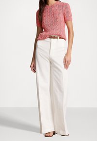 Pull en maille torsadée rose à manches courtes avec col rond, associé à un pantalon large taille haute blanc et une ceinture tressée. Sandales plates visibles.