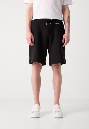 Persoon die zwarte shorts met trekkoord, witte sneakers en een wit T-shirt draagt, staande tegen een effen lichte achtergrond.