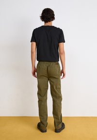 Black short-sleeve t-shirt and olive green vintage-style pants with an elastic waistband, viewed from the back; black shoes with yellow stitching.